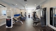 This fitness center is organized into distinct functional zones, offering a comprehensive workout environment within a single-story layout. The space is finished with durable dark rubber flooring throughout, providing traction and impact absorption suitable for a wide range of exercises.

The cardio area houses multiple treadmills positioned near large windows that invite natural light and outdoor views of surrounding greenery. A ceiling fan overhead aids air circulation, while a wall-mounted flat-screen TV keeps users engaged during workouts. A bold blue accent stripe along the wall adds a dynamic visual element to this zone.

The strength training area is well-appointed with a multi-station cable machine, adjustable benches, and a full rack of dumbbells neatly organized on a tiered stand. Mirrors and additional wall-mounted TVs are positioned throughout, enhancing both functionality and the sense of spaciousness.

A dedicated functional training nook features resistance bands, stability balls, step platforms, and a flat-screen display for guided workouts. An arched window in this area provides a bright, inviting atmosphere. Recessed lighting and ceiling fans are distributed across the facility, ensuring consistent illumination and comfort throughout.