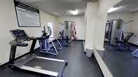 This community fitness room is a dedicated exercise space designed exclusively for residents. The room features a dark, durable floor surface well-suited for a high-traffic workout environment, with neutral-toned walls and overhead fluorescent lighting that keep the space bright and functional.

The fitness room is equipped with a range of exercise machines to support both cardiovascular and strength training. Cardio options include a treadmill and an elliptical trainer, offering residents versatile options for aerobic workouts. For strength training, a multi-function cable and weight machine is positioned toward the back of the room, complemented by an adjustable workout bench. A large wall-mounted mirror along one side of the room enhances the sense of space and allows users to monitor their form during exercise.

Additional amenities include waste bins conveniently placed near the mirror wall, and a small shelf or ledge providing a surface for personal items. A posted set of fitness room rules is visible near the entrance, reinforcing community guidelines for safe and respectful use of the space. A window allows for a degree of natural light. The layout is practical and efficiently organized, making the most of the available space to provide residents with a well-rounded, accessible fitness facility.