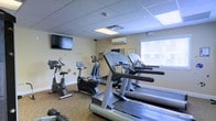 This single-floor fitness room provides a practical and functional workout environment designed to meet everyday exercise needs. The space is outfitted with a selection of cardio equipment, including multiple treadmills, an elliptical machine, and stationary bikes, offering users a variety of training options. A wall-mounted flat-screen television is positioned at eye level, allowing occupants to stay entertained during workouts. A split-system air conditioning unit ensures the room remains comfortable regardless of the season. Natural light enters through a window fitted with horizontal blinds, contributing to a bright and pleasant atmosphere. The drop ceiling with recessed and flush-mounted light panels provides consistent, even illumination throughout the space. The floor is covered with a patterned carpet, adding a degree of softness underfoot. The walls are painted in a neutral tone, keeping the room feeling open and clean. Small decorative elements, including colorful wall decals, add a subtle touch of personality to the otherwise utilitarian setting. A wall mirror is also present, a practical feature for monitoring form during exercise.