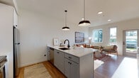 Located at 7900 SE Luther Rd in Portland, Oregon, this thoughtfully designed single-floor residence offers a clean, modern aesthetic with a warm and inviting ambiance. The open-concept layout seamlessly connects the kitchen and living area, creating a cohesive and functional living space. The kitchen is a standout feature, equipped with a central island topped with white quartz countertops, gray lower cabinetry, white upper cabinetry, stainless steel appliances including a range, microwave, and refrigerator, and a black matte undermount sink. Stylish black pendant lights hang above the island, adding a contemporary accent. Light hardwood flooring runs throughout the unit, enhancing the bright and airy feel. The living area is generously proportioned, benefiting from multiple windows that invite abundant natural light and provide views of the surrounding neighborhood. Two bedrooms offer comfortable accommodations, each with hardwood floors and good natural light, one featuring large windows with outdoor views. The full bathroom is well-appointed with a dark navy vanity, quartz countertop, undermount sink, a large mirror, and a full tub-shower combination. The overall design balances modern minimalism with functional, livable details throughout.