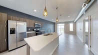 This single-floor space showcases a thoughtfully designed open-concept layout that seamlessly connects the kitchen and living area. The kitchen is a standout feature, anchored by a large central island with a sleek white quartz countertop and an undermount double sink. Stainless steel appliances, including a refrigerator, electric range, and over-the-range microwave, are set against wood-toned cabinetry with modern hardware and a classic tile backsplash. A muted blue accent wall adds depth and character to the kitchen space. Two eye-catching gold teardrop pendant lights hang above the island, providing warm, focused illumination that complements the recessed ceiling lighting throughout. The flooring transitions smoothly from the kitchen into the open living area, finished in light-toned hardwood-style planks that enhance the sense of space and continuity. The living area is bright and airy, benefiting from large windows that flood the interior with natural light and offer open outdoor views. The overall ambiance is clean, contemporary, and inviting, with a neutral palette punctuated by warm metallic accents and modern design details.