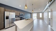 This single-floor space showcases a thoughtfully designed open-concept layout that seamlessly connects the kitchen and living area. The kitchen is a standout feature, anchored by a large central island with a sleek white quartz countertop and an undermount double sink. Stainless steel appliances, including a refrigerator, electric range, and over-the-range microwave, are set against wood-toned cabinetry with modern hardware and a classic tile backsplash. A muted blue accent wall adds depth and character to the kitchen space. Two eye-catching gold teardrop pendant lights hang above the island, providing warm, focused illumination that complements the recessed ceiling lighting throughout. The flooring transitions smoothly from the kitchen into the open living area, finished in light-toned hardwood-style planks that enhance the sense of space and continuity. The living area is bright and airy, benefiting from large windows that flood the interior with natural light and offer open outdoor views. The overall ambiance is clean, contemporary, and inviting, with a neutral palette punctuated by warm metallic accents and modern design details.