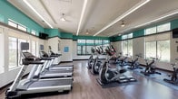 This fitness center is a thoughtfully designed, single-floor facility built to support a wide range of workout routines. The space features a bold color scheme of teal accent walls paired with neutral whites and charcoal trim, creating an energizing atmosphere throughout. Durable wood-look flooring runs across the entire facility, complemented by recessed linear lighting and ceiling fans that ensure the space remains well-lit and comfortable during exercise.

The main workout floor is generously sized and outfitted with a comprehensive selection of cardio equipment, including multiple treadmills, elliptical trainers, and stationary cycling bikes arranged in organized rows. Large windows along the perimeter flood the space with natural light and offer views of the surrounding greenery. Wall-mounted televisions are positioned throughout for entertainment during workouts, and full-length mirrors expand the visual depth of the room.

A dedicated strength and sports training area is enclosed by floor-to-ceiling glass panels, providing visibility into a space equipped with weight racks, barbells, and turf flooring designed for functional training. Near the entrance, a service counter with built-in storage offers space for sanitizing supplies and towels, keeping the facility clean and organized. The overall layout balances openness with purpose, making this fitness center a fully functional and inviting space for all fitness levels.