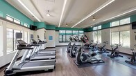 This fitness center is a thoughtfully designed, single-floor facility built to support a wide range of workout routines. The space features a bold color scheme of teal accent walls paired with neutral whites and charcoal trim, creating an energizing atmosphere throughout. Durable wood-look flooring runs across the entire facility, complemented by recessed linear lighting and ceiling fans that ensure the space remains well-lit and comfortable during exercise.

The main workout floor is generously sized and outfitted with a comprehensive selection of cardio equipment, including multiple treadmills, elliptical trainers, and stationary cycling bikes arranged in organized rows. Large windows along the perimeter flood the space with natural light and offer views of the surrounding greenery. Wall-mounted televisions are positioned throughout for entertainment during workouts, and full-length mirrors expand the visual depth of the room.

A dedicated strength and sports training area is enclosed by floor-to-ceiling glass panels, providing visibility into a space equipped with weight racks, barbells, and turf flooring designed for functional training. Near the entrance, a service counter with built-in storage offers space for sanitizing supplies and towels, keeping the facility clean and organized. The overall layout balances openness with purpose, making this fitness center a fully functional and inviting space for all fitness levels.