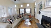 This traditionally styled single-floor space exudes warmth and classic charm throughout. The main living area immediately draws the eye upward with its impressively tall ceilings, accentuated by a row of clerestory windows positioned high on the walls that flood the room with natural light alongside the larger standard windows below. Rich hardwood flooring runs throughout, anchoring the space with a sense of timeless elegance.

The living room is generously proportioned, comfortably accommodating a full suite of classic upholstered furnishings, including floral-patterned sofas and loveseats arranged around a solid wood coffee table — reflecting a cozy, lived-in aesthetic. Decorative drapes with bold floral patterns frame the windows, complementing the warm, neutral tones of the walls finished with crisp crown molding details.

A built-in white cabinetry unit adds both functionality and architectural interest to one side of the room, while an arched entryway provides a graceful transition between spaces. The overall ambiance is inviting and comfortable, blending traditional design elements — from ornate light fixtures to carefully curated décor — to create a space that feels both distinguished and approachable.