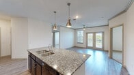 Located at 1803 W Ennis Ave in Ennis, Texas, this well-appointed 2-bedroom, 2-bathroom single-story home offers a thoughtfully designed layout with modern finishes throughout. The open-concept kitchen serves as the heart of the home, showcasing dark espresso cabinetry, granite countertops, a center island with a built-in sink, and stylish pendant lighting overhead. Black appliances including a refrigerator, range, and microwave complete the functional kitchen setup. Wood-look luxury vinyl plank flooring runs throughout the main living areas, adding warmth and continuity to the space. The living room is bright and inviting, featuring crown molding, a ceiling fan, and French doors that open to a private patio, allowing for natural light and outdoor views. Both bedrooms are carpeted for comfort, with the primary bedroom offering a spacious walk-in closet equipped with wire shelving systems. The primary bathroom features a dual-sink vanity with granite countertops, dark cabinetry, and a walk-in shower. The second bedroom is served by a separate full bathroom. Additional highlights include a dedicated laundry area, entrance foyer, hallway, and four closets throughout, ensuring ample storage. The home's clean, neutral palette and modern design elements create a cohesive and move-in-ready atmosphere.