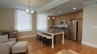 This single-floor space showcases an open-concept layout that seamlessly integrates the kitchen and living areas, creating a cohesive and functional environment. The kitchen is a standout feature, equipped with stainless steel appliances including a refrigerator, range, and built-in microwave. Rich medium-toned wood cabinetry lines the walls, complemented by dark granite countertops that add depth and elegance to the space.

A large central island with a contrasting white base and matching granite surface serves as the focal point of the kitchen, offering bar-style seating with several stools — ideal for casual dining or entertaining. Pendant lighting above the island adds warm, ambient illumination, while a classic chandelier near the dining area enhances the overall aesthetic.

Warm hardwood floors run throughout, contributing to the inviting atmosphere of the space. Large windows dressed with white blinds allow natural light to fill the room, brightening the neutral-toned walls. Crown molding along the ceiling adds a refined architectural touch, and recessed lighting ensures consistent illumination throughout the open living and kitchen areas.