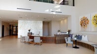 This expansive lobby presents a striking contemporary design anchored by soaring double-height ceilings and a dramatic floor-to-ceiling glass façade that floods the interior with natural light and provides views of mature palm trees beyond. Large-format light-toned porcelain tiles extend throughout, reinforcing the open, airy atmosphere.

The main reception area features a custom curved desk with warm wood slatted detailing, set against a commanding backlit onyx or marble stone wall feature that serves as a focal point. A secondary service counter nearby offers additional functionality. Curated original artwork is displayed throughout, including vibrant floral canvases and abstract triptychs, contributing a gallery-like quality to the space.

Intimate seating alcoves are positioned throughout, furnished with upholstered lounge chairs, accent tables, and framed views of the landscaped exterior. A dedicated mail and package room includes an extended communal table and organized parcel lockers. The elevator lobby features a luminous backlit stone surround with bronze-finished elevator doors and decorative mosaic floor tile detailing. Thoughtfully placed greenery and pendant lighting complete the polished, welcoming ambiance.