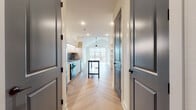 This thoughtfully designed single-floor residence showcases a clean, contemporary aesthetic throughout. The open-concept living area is anchored by soaring vaulted ceilings and a ceiling fan, creating an airy, expansive atmosphere. A large, multi-panel window wall floods the space with natural light and provides direct access to an outdoor balcony, offering views of the surrounding neighborhood. The herringbone-patterned hardwood flooring flows seamlessly through every room, adding warmth and visual texture to the modern palette of crisp white walls and dark hardware accents.

The kitchen is outfitted with flat-front cabinetry in a warm neutral tone, under-cabinet LED lighting, a sleek gray tile backsplash, an induction cooktop, a built-in oven, stainless steel appliances, and a dark-topped island that serves as both a workspace and gathering point. A corridor lined with dark panel doors and built-in open shelving connects the main living area to the private spaces.

The bedroom is a serene retreat with the same herringbone flooring and a ceiling fan overhead. It is accompanied by a generously sized walk-in closet featuring multi-level hanging rods and dedicated shelving. The bathroom offers a refined finish with a bold, floor-to-ceiling blue tile walk-in shower with a frameless glass enclosure, a floating vanity, oval LED mirrors, and built-in shelving for added storage.