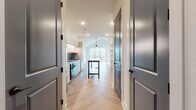 This thoughtfully designed single-floor residence showcases a clean, contemporary aesthetic throughout. The open-concept living area is anchored by soaring vaulted ceilings and a ceiling fan, creating an airy, expansive atmosphere. A large, multi-panel window wall floods the space with natural light and provides direct access to an outdoor balcony, offering views of the surrounding neighborhood. The herringbone-patterned hardwood flooring flows seamlessly through every room, adding warmth and visual texture to the modern palette of crisp white walls and dark hardware accents.

The kitchen is outfitted with flat-front cabinetry in a warm neutral tone, under-cabinet LED lighting, a sleek gray tile backsplash, an induction cooktop, a built-in oven, stainless steel appliances, and a dark-topped island that serves as both a workspace and gathering point. A corridor lined with dark panel doors and built-in open shelving connects the main living area to the private spaces.

The bedroom is a serene retreat with the same herringbone flooring and a ceiling fan overhead. It is accompanied by a generously sized walk-in closet featuring multi-level hanging rods and dedicated shelving. The bathroom offers a refined finish with a bold, floor-to-ceiling blue tile walk-in shower with a frameless glass enclosure, a floating vanity, oval LED mirrors, and built-in shelving for added storage.