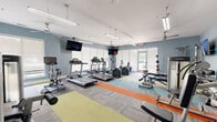 This fitness center offers a comprehensive workout environment housed within a single open-plan space. The room is finished with light blue walls and a crisp white ceiling, lending a clean and energizing atmosphere. Overhead, a combination of fluorescent strip lighting and ceiling fans ensures the space remains well-lit and comfortable during exercise.

The flooring is a standout design feature, composed of modular carpet tiles arranged in bold, colorful stripes of yellow-green, orange, teal, and gray, delineating different workout zones throughout the room.

The cardio zone is prominently equipped with multiple treadmills, elliptical trainers, stationary bikes, and a recumbent bike, all arranged to face large windows fitted with roller shades that diffuse natural light into the space. Multiple wall-mounted flat-screen televisions are positioned throughout the room, providing entertainment during workouts.

The strength training area includes a cable machine, adjustable weight bench, dumbbell rack, medicine balls, and a variety of resistance and functional training equipment. A mirrored section along one wall enhances the sense of space and supports proper form during training.

The fitness center is accessible via both a set of French doors and a direct exterior entrance, offering convenient access from multiple points.