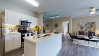 This single-floor residence is thoughtfully designed with a cohesive, contemporary aesthetic that flows naturally from room to room. The open-concept kitchen serves as a central hub, featuring a large white peninsula with an integrated sink and gold-toned faucet, crisp white cabinetry, a subway tile backsplash, and a full suite of stainless steel appliances including a refrigerator, range, and over-the-range microwave. Wood-look flooring extends throughout the main living areas, adding warmth and continuity.

The living room is generously proportioned, finished with crown molding and neutral-toned walls that create a calm, welcoming atmosphere. A sliding glass door with vertical blinds fills the space with natural light and opens to a private balcony. Track lighting in the transitional areas provides additional illumination.

Two bedrooms offer plush carpeting, ceiling fans, and accent walls in a deep charcoal tone that contrast stylishly against the neutral surroundings. Each room accommodates a large bed comfortably, with windows providing natural light. The bathroom includes a white vanity with a granite countertop, under-cabinet storage, and a shower-tub combination.