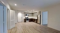 This single-floor space showcases a thoughtfully designed open-concept layout that seamlessly blends functionality with modern aesthetics. The kitchen serves as a central highlight, featuring a full suite of stainless steel appliances including a gas range with oven, built-in microwave, dishwasher, and a full-size refrigerator. Cabinetry is finished in a two-tone style, combining upper light gray cabinets with rich dark wood lower cabinets, creating a striking and contemporary contrast. Light-toned countertops complement the palette, while a recessed sink completes the kitchen workspace. Recessed lighting throughout the ceiling provides bright, even illumination across the entire space. The floors are finished with light wood-toned luxury vinyl planks, contributing to an airy and welcoming ambiance. The open floor plan allows the kitchen to flow naturally into the surrounding living area, offering a spacious feel and flexible use of the space. Doors with clean white trim are visible leading to additional areas of the unit. The overall design reflects a modern, move-in-ready aesthetic with neutral tones and quality finishes that appeal to a wide range of tastes.