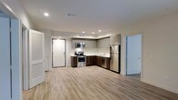 This single-floor space showcases a thoughtfully designed open-concept layout that seamlessly blends functionality with modern aesthetics. The kitchen serves as a central highlight, featuring a full suite of stainless steel appliances including a gas range with oven, built-in microwave, dishwasher, and a full-size refrigerator. Cabinetry is finished in a two-tone style, combining upper light gray cabinets with rich dark wood lower cabinets, creating a striking and contemporary contrast. Light-toned countertops complement the palette, while a recessed sink completes the kitchen workspace. Recessed lighting throughout the ceiling provides bright, even illumination across the entire space. The floors are finished with light wood-toned luxury vinyl planks, contributing to an airy and welcoming ambiance. The open floor plan allows the kitchen to flow naturally into the surrounding living area, offering a spacious feel and flexible use of the space. Doors with clean white trim are visible leading to additional areas of the unit. The overall design reflects a modern, move-in-ready aesthetic with neutral tones and quality finishes that appeal to a wide range of tastes.