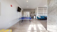 Located at 200 Carpenter St in Warrensburg, Missouri, this single-floor game room is a thoughtfully designed recreational space built for socializing and entertainment. The room is finished with light-toned large-format tile flooring throughout, contributing to a clean, modern aesthetic that is both stylish and easy to maintain.

At the heart of the space is a striking pool table with a bold blue felt surface, positioned beneath a ceiling fan and complemented by a textured decorative wall panel. A full-size ping-pong table provides additional entertainment options. Comfortable modern seating — including a teal sectional sofa and accent chairs — is arranged to encourage lounging between games.

A standout architectural feature is the floor-to-ceiling marble-clad column and fireplace wall, which anchors the room with an air of sophistication. A wall-mounted flat-screen TV is positioned above the fireplace, creating an ideal entertainment focal point. Additional mounted TVs are positioned throughout the space for versatile viewing.

Natural light pours in through large windows and glass-paneled French doors, keeping the space bright and open. A small kitchenette with open shelving and a compact refrigerator is tucked into one corner, adding everyday convenience. A bulletin board near the entrance provides a practical communal touch, rounding out this versatile and inviting gathering space.