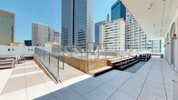 The Mayflower Pool is a rooftop amenity that delivers an elevated outdoor experience set against a striking downtown skyline backdrop. The centerpiece is a generously sized swimming pool finished with mosaic tile coping, offering shallow sun ledges ideal for lounging. Surrounding the pool, a broad wooden deck is outfitted with rows of contemporary chaise lounges and low-profile side tables, providing ample space for sunbathing and socializing.

A tiered wooden platform area with stainless steel railings creates distinct lounging zones at varying levels, adding architectural interest to the deck layout. Glass perimeter railings along the outer edges preserve unobstructed panoramic views of the surrounding high-rise skyline, visible from nearly every point on the terrace.

An outdoor kitchen area features dual stainless steel grills set against a dark brick backsplash, with ample counter space for outdoor dining and entertaining. A covered pavilion structure with arched openings provides shaded seating areas directly adjacent to the pool.

Large-format tile paving transitions seamlessly to the warm-toned wood decking throughout the terrace, creating a cohesive aesthetic. Access is provided through floor-to-ceiling sliding glass doors, clearly marked as a resident pool. The space is bathed in natural sunlight, making it a vibrant urban retreat above the city.