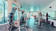 This fitness center in Miami, FL offers a bright, well-organized exercise room designed to support a comprehensive workout experience. The space features durable rubber flooring throughout, providing a safe and functional surface for a wide range of activities. Large windows line the walls, flooding the room with natural light and offering views of lush tropical palm trees — a defining characteristic of the Miami setting. Glass doors at one end provide direct access to the outdoors, further enhancing the open, airy atmosphere.

The exercise room is equipped with a variety of cardio machines, including multiple treadmills and elliptical trainers, as well as strength training equipment such as a multi-station cable machine, a Smith machine with an adjustable bench, and free weights. Full-length mirrors are mounted along one wall, supporting proper form during workouts and visually expanding the space.

Structural columns are integrated into the layout, complementing the clean white walls and vaulted ceiling sections that add architectural interest. Recessed and flush-mounted lighting ensures the space is well-lit at all hours. The overall ambiance is functional and inviting, combining practical fitness amenities with the bright, tropical character of its Miami location.
