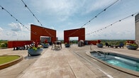 Located at 7475 Flying Cloud Dr in Eden Prairie, Minnesota, this two-level property features a standout rooftop amenity deck that delivers an exceptional outdoor experience. The expansive rooftop terrace is finished with large-format stone pavers and anchored by prominent red brick architectural walls that frame the space and add a bold, industrial character.

At the center of the deck sits a built-in hot tub, accessible via stainless steel entry rails and surrounded by open terrace space ideal for relaxation. An outdoor kitchen area features a built-in grill station with a stacked stone surround, offering a dedicated space for outdoor cooking and entertaining. Multiple bar-height table and stool sets are arranged throughout the terrace, providing ample seating for gatherings. A putting green adds a recreational element to the layout.

Large decorative planters filled with vibrant seasonal flowers and ornamental grasses are positioned throughout the deck, bringing color and life to the space. Overhead string lights are strung across the entire terrace, creating an inviting ambiance for evening use.

The rooftop commands sweeping panoramic views of the surrounding Eden Prairie landscape, including the treeline and neighboring developments. A yellow-sided amenity building with direct rooftop access provides shelter and support facilities. The open sky and abundant natural light make this rooftop an engaging and versatile outdoor destination.
