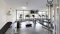 Located at 30 Esterbrooke Avenue in Toronto's M2J neighbourhood, this fitness room offers a well-equipped exercise space on a single level. The room is thoughtfully organized to maximize its footprint, accommodating a variety of workout options within a compact yet functional layout.

The cardio section is positioned along a wall of large floor-to-ceiling windows that flood the space with natural light and provide pleasant views of the surrounding greenery and neighbourhood skyline — an inviting backdrop for a workout. Equipment includes a Life Fitness treadmill, an elliptical trainer, and a stationary bike. A wall-mounted flat-screen television is installed above the windows, adding convenience for users during cardio sessions.

A full-length mirror spans one wall, visually expanding the room and allowing users to monitor their form. The strength-training area features a Life Fitness dual cable machine with suspension attachments, a dumbbell rack with a range of free weights, an adjustable weight bench, and a stability ball. The durable rubber flooring throughout provides excellent traction and cushioning. Ceiling-mounted flush lighting complements the natural light. The room is accessed through a private door and maintains a clean, neutral aesthetic with white walls and a bright, open ambiance suited for focused training.