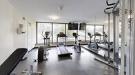 Located at 30 Esterbrooke Avenue in Toronto's M2J neighbourhood, this fitness room offers a well-equipped exercise space on a single level. The room is thoughtfully organized to maximize its footprint, accommodating a variety of workout options within a compact yet functional layout.

The cardio section is positioned along a wall of large floor-to-ceiling windows that flood the space with natural light and provide pleasant views of the surrounding greenery and neighbourhood skyline — an inviting backdrop for a workout. Equipment includes a Life Fitness treadmill, an elliptical trainer, and a stationary bike. A wall-mounted flat-screen television is installed above the windows, adding convenience for users during cardio sessions.

A full-length mirror spans one wall, visually expanding the room and allowing users to monitor their form. The strength-training area features a Life Fitness dual cable machine with suspension attachments, a dumbbell rack with a range of free weights, an adjustable weight bench, and a stability ball. The durable rubber flooring throughout provides excellent traction and cushioning. Ceiling-mounted flush lighting complements the natural light. The room is accessed through a private door and maintains a clean, neutral aesthetic with white walls and a bright, open ambiance suited for focused training.