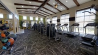 This fitness center offers a thoughtfully designed single-floor workout facility that combines functionality with an inviting aesthetic. The space is anchored by striking exposed dark wood ceiling beams set against a crisp white vaulted ceiling, lending the room a warm yet airy character. Ceiling fans are positioned throughout to maintain comfortable airflow during workouts.

Large windows line the walls, flooding the interior with natural light and providing views to the outdoors, creating an open and energizing atmosphere. Wall-mounted flat-screen TVs are conveniently positioned throughout the space for entertainment during workouts.

The floor is covered in a boldly patterned carpet featuring an abstract swirling design in dark tones with light accents, adding a dynamic visual element to the space. Mirrors along one wall enhance the sense of depth and allow users to monitor their form effectively.

The facility is outfitted with a comprehensive array of modern exercise equipment, including multiple treadmills, a full selection of strength-training cable and selectorized machines, and additional cardio options. Accent walls in a vibrant yellow-green add a lively pop of color, complementing the neutral tones of the walls and contributing to the motivating environment this fitness center provides.