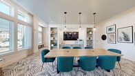 Located in New Port Richey, Florida, this thoughtfully designed single-floor community space offers a versatile and stylish environment suited for meetings, collaboration, and relaxation. The interior is anchored by a bold geometric-patterned carpet that runs throughout, complementing the clean white walls and bright, airy atmosphere.

The main gathering area features a large conference table surrounded by teal upholstered chairs, pendant lighting overhead, and a wall-mounted flat-screen TV flanked by built-in shelving units — ideal for presentations or group discussions. A printer station and decorative accessories are neatly integrated into the built-ins, adding both function and visual interest.

Adjacent to the conference area, glass-paneled French doors lead to a private enclosed meeting room, offering a quieter setting for smaller gatherings. The open layout transitions seamlessly into a comfortable lounge zone, where cushioned armchairs and side tables are arranged beneath floor-to-ceiling windows dressed with sheer drapes and horizontal blinds, filling the space with generous natural light and outdoor views.

A curated gallery wall featuring framed botanical prints and circular black accents adds a modern artistic touch. Built-in wall niches with decorative shelving provide additional visual depth. The overall ambiance is polished, welcoming, and professionally appointed.