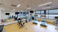 This fitness center offers a comprehensive workout environment designed to accommodate a wide range of exercise needs. The space is laid out on a single floor with cork flooring throughout, providing a resilient and comfortable surface underfoot. A suspended drop ceiling with recessed and linear fluorescent lighting ensures the area is uniformly and brightly illuminated.

The cardio zone is anchored by multiple treadmills, elliptical trainers, and a stationary bike, all positioned along the exterior windows. These large windows invite natural light and offer outdoor views, including access to a furnished outdoor terrace through a glass door — ideal for fresh-air breaks. Wall-mounted flat-screen televisions are strategically placed for entertainment during workouts.

A full-length mirror wall visually expands the space and is complemented by a ballet-style barre and resistance band attachments, supporting stretching and flexibility routines. A dumbbell rack with a full set of free weights, along with a water cooler station, adds functional convenience. Stability balls and additional strength training equipment round out the offerings.

Along one wall, built-in cabinetry in a light wood finish provides organized storage, while framed motivational artwork decorates the sage-green accent walls. The facility is noted as available 24 hours, making it a highly accessible and versatile fitness destination.