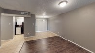 This single-floor unit offers a practical and straightforward layout with well-defined living spaces throughout. The living area features a combination of dark hardwood-style flooring and light ceramic tile, set against neutral gray walls and a textured ceiling. A large window allows natural light to filter in, brightening the open space. The adjoining dining area continues the tile flooring and is illuminated by a pendant light fixture, with vertical blinds covering a sliding glass door.

The kitchen is equipped with black appliances, including a freestanding electric range, a refrigerator, and a ventilation hood. White cabinetry provides ample storage above and below, complemented by speckled countertops and a dark mosaic tile backsplash that adds a subtle design accent.

The bedroom features light tile flooring, gray walls, a textured ceiling, and access to a closet, offering a clean and functional space. A second room with similar finishes and a window provides additional flexible living space.

The bathroom includes a full tub and shower combination with large-format beige ceramic tile surrounds, a white toilet, and a granite-style countertop vanity with dual sinks, all finished with chrome fixtures. Throughout the unit, white trim and baseboards provide a clean contrast to the gray walls.