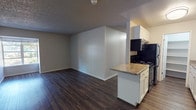 This single-floor residence offers a practical and comfortable layout designed for everyday living. The open-concept main area seamlessly connects the kitchen and living space, both finished with rich, dark hardwood-style flooring that adds warmth and visual continuity. The kitchen is well-equipped with white cabinetry, a peninsula counter with a laminate surface, black appliances including a gas range and microwave, a stainless steel dishwasher, and a sink with ample counter space. A textured ceiling runs throughout the unit, adding a classic touch to the interior. The living area is generously sized with baseboard heating along the wall, ensuring year-round comfort. Two bedrooms are carpeted for a softer underfoot feel and each features a window overlooking mature trees, allowing for natural light and pleasant outdoor views. Both rooms are served by baseboard heating as well. The full bathroom is clean and functional, featuring a white vanity with a laminate countertop, a large mirror with overhead lighting, and a shower-tub combination. The entry door opens directly into the main living and kitchen area, creating an efficient and straightforward floor plan suited for comfortable, practical living.