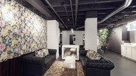 This single-floor space makes a bold visual statement through its thoughtful contrast of industrial structure and vibrant, curated interior design. The exposed ceiling features raw black-painted beams and structural elements, lending an urban, warehouse-inspired character to the space. Track lighting is integrated throughout, providing focused illumination across the open layout.

White structural columns anchor the floor plan, defining zones within the open-concept interior. At the heart of the space sits a freestanding double-sided fireplace with a sleek white surround, serving as a striking focal point. Surrounding it, tufted black leather sofas are arranged to create an intimate and stylish lounge setting, complemented by a marble-topped coffee table.

Underfoot, a richly patterned area rug in warm, multicolored tones adds texture and warmth to the space. One wall is adorned with bold floral wallpaper in pastel tones — pink, yellow, green, and blue — that introduces color and artistic flair. Matching floral-print throw pillows echo this motif across the seating area. Toward the perimeter, lush greenery adds a natural, organic contrast to the industrial ceiling above. The overall ambiance is sophisticated yet energetic, blending comfort with creative design sensibility.
