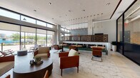 This thoughtfully designed single-floor space combines a welcoming lounge, a dining area, and a self-serve market into one cohesive environment. The interior is anchored by large floor-to-ceiling windows along the perimeter, flooding the space with abundant natural light and offering views of the surrounding outdoor streetscape. Light wood-plank flooring and large-format hexagonal stone-look tile unite the various zones seamlessly.

The main lounge area features a curated mix of mid-century modern seating, including cognac leather armchairs with brass-accented frames and boucle upholstered chairs arranged around low, rounded wood tables. Recessed ceiling lighting and a striking row of cascading brass pendant lights add warmth and visual interest overhead. A decorative laser-cut wood screen partition separates the lounge from a raised banquette dining zone, which offers plush green upholstered bench seating alongside intimate café tables.

A secondary dining area near the exterior entrance includes upholstered booth seating, oval dining tables with leather chairs, and woven rattan pendant lighting, reinforcing the space's warm, organic aesthetic. Adjacent to this is a casual market nook enclosed by glass partitions, furnished with high-top tables, bar stools, and refrigerated display cases stocked with beverages and snacks. Lush potted greenery is scattered throughout, enhancing the inviting, resort-like ambiance.