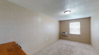 Located at 12204 NW Heady Ave in Ferrelview, Missouri, this single-floor space offers a straightforward and functional layout designed for comfortable everyday living. The main living area is finished with soft wall-to-wall carpeting and textured walls, creating a warm and neutral aesthetic throughout. A wall-mounted air conditioning unit provides climate control, while a window with blinds invites natural light and outdoor views into the room. The textured ceiling adds a classic character consistent across the space. A dedicated kitchenette nook features a wall-mounted wooden shelf and a compact refrigerator, offering a practical setup for small-scale food and beverage storage. The bathroom area is a standout feature, showcasing a spacious layout with terracotta-toned tile flooring, an extensive built-in shelving and closet rod system for generous storage, and a glass-door walk-in shower enclosure. A dark wood-toned door adds a contrasting design element in this area. The overall ambiance of the space is clean, neutral, and move-in ready, with each area thoughtfully organized to maximize utility within the available footprint.