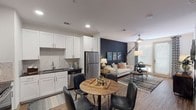 This single-floor residence offers a well-designed open-concept layout that seamlessly connects the kitchen, dining, and living areas. The kitchen is outfitted with white shaker-style cabinetry, dark countertops, a herringbone tile backsplash, and a full suite of stainless steel appliances, including a refrigerator, dishwasher, range, and built-in microwave. Wood-look flooring runs throughout, providing a warm, cohesive aesthetic. The dining area accommodates a round table with seating for four and flows naturally into the living room, which features a bold navy accent wall, a ceiling fan, and a glass door that welcomes natural light. The bedroom is highlighted by a striking black-and-white striped accent wall, a ceiling fan, and ample room for a full furniture arrangement. A dedicated den or office area provides a quiet, versatile space suitable for working from home or relaxing. The bathroom includes a full soaking tub with a white subway tile surround, open shelving for storage, and wood-look flooring that continues the design language of the rest of the unit. A walk-in closet with multi-tier shelving is conveniently accessible from the bathroom, offering generous storage capacity throughout this thoughtfully appointed residence.