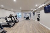 Located on Cedar St N in Kitchener, Ontario, this fitness facility offers a generously sized, single-floor gym designed to accommodate a full range of workout needs. The space features a clean, modern aesthetic with neutral-toned walls, light wood-style flooring, and recessed ceiling lighting throughout, creating a bright and welcoming environment conducive to exercise.

The main gym area is thoughtfully organized into distinct workout zones. A dedicated cardio section houses multiple treadmills and stationary cycling bikes, providing ample capacity for simultaneous users. A separate area accommodates several elliptical cross-trainers, offering low-impact cardiovascular options. Strength training is well-served by a multi-station cable and weight machine setup, suitable for a wide variety of resistance exercises.

Structural columns divide the open floor plan into natural activity zones without compromising the overall sense of spaciousness. The flooring throughout is easy-to-maintain and durable, appropriate for high-traffic fitness use. Decorative artwork is placed on the walls, adding a polished, finished quality to the space. A wall-mounted television screen is also present, enhancing the workout experience. Access points include a key-fob secured entry door, and safety signage is clearly posted. The overall design balances functionality with a clean, contemporary aesthetic.