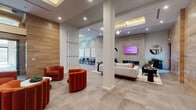 This thoughtfully designed single-floor amenity space blends contemporary aesthetics with functional versatility across several well-defined zones. The entry foyer welcomes visitors with large-scale black-framed glass doors and clerestory windows that draw in natural light, complemented by a coffered ceiling with warm wood inlay detailing. Bold terracotta-orange velvet lounge chairs arranged around a round marble-topped table create an inviting focal point near the entrance.

The main lounge area transitions seamlessly from the entry, anchored by a floor-to-ceiling stone tile fireplace wall with a wall-mounted flat-screen television and flanked by round decorative mirrors. Plush neutral sofas, sculptural accent chairs, and curated greenery contribute to a relaxed yet refined atmosphere throughout.

A dedicated co-working and social kitchen area features a substantial marble-topped island with seating, warm wood cabinetry, a built-in sink, and direct access to an outdoor seating area through full-height glass sliding doors. Wood-paneled walls add warmth and continuity.

Private conference or focus rooms are enclosed with glass partitions, offering visual connectivity while maintaining acoustic separation. One features textured dark wall tile, a long built-in white desk, and curated abstract artwork, creating a refined workspace. Large-format stone tile flooring unifies the entire space.