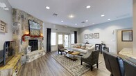 This single-floor space offers a thoughtfully designed interior that balances comfort and style. The living area serves as the focal point, anchored by a striking stone fireplace that extends to the ceiling and is complemented by a mounted flat-screen television above the mantle. The fireplace's natural stonework adds a rustic yet refined character to the space.

Hardwood-style flooring runs throughout, providing a warm, cohesive foundation. Recessed lighting is installed across the ceiling, offering consistent, well-distributed illumination that enhances the room's brightness alongside natural light from multiple windows and French doors. The French doors open to an outdoor area, allowing for a seamless indoor-outdoor connection.

The seating arrangement includes a neutral-toned sofa with colorful accent pillows, wicker-style armchairs, and a wooden coffee table, all positioned atop a patterned area rug. On the opposite wall, a small console table is paired with framed artwork, adding a decorative touch. The soft gray wall tones, combined with the warm flooring and natural elements, create an overall ambiance that feels both polished and inviting.