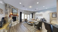 This single-floor space offers a thoughtfully designed interior that balances comfort and style. The living area serves as the focal point, anchored by a striking stone fireplace that extends to the ceiling and is complemented by a mounted flat-screen television above the mantle. The fireplace's natural stonework adds a rustic yet refined character to the space.

Hardwood-style flooring runs throughout, providing a warm, cohesive foundation. Recessed lighting is installed across the ceiling, offering consistent, well-distributed illumination that enhances the room's brightness alongside natural light from multiple windows and French doors. The French doors open to an outdoor area, allowing for a seamless indoor-outdoor connection.

The seating arrangement includes a neutral-toned sofa with colorful accent pillows, wicker-style armchairs, and a wooden coffee table, all positioned atop a patterned area rug. On the opposite wall, a small console table is paired with framed artwork, adding a decorative touch. The soft gray wall tones, combined with the warm flooring and natural elements, create an overall ambiance that feels both polished and inviting.
