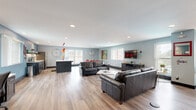 Located at 3208 W Seventh St in Greeley, Colorado, this single-floor community space offers a thoughtfully designed, open-concept layout that seamlessly integrates multiple functional zones. The interior is finished with durable wood-look flooring throughout and painted in a calming blue-gray palette that lends the space a welcoming, contemporary feel.

The expansive lounge area is a central feature, furnished with multiple leather sofas arranged around a sleek coffee table and a wall-mounted flat-screen TV. Multiple windows allow natural light to fill the room, creating a bright and comfortable environment. The space accommodates both relaxed gathering and structured activities with ease.

The kitchen is modern and well-appointed, featuring dark cabinetry, white quartz countertops, stainless steel appliances including a full-size refrigerator and dishwasher, and a large island with bar seating. Striking red pendant lights hang above the island, adding a bold design accent. Open shelving provides convenient storage and display space.

A dedicated workstation area along one wall is equipped with desks, computers, and office chairs, supporting productivity and administrative needs. A small dining area with a table and chairs offers an additional gathering spot. Recessed lighting throughout ensures the entire space is well-lit. The overall design balances comfort, functionality, and community-oriented use.