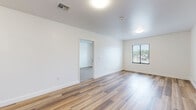 Located at 1120 S Walton Blvd in Bentonville, Arkansas, this single-floor unit offers a clean, modern layout designed for functional everyday living. The main living area is expansive and open, featuring warm wood-look laminate flooring that extends throughout the non-carpeted spaces, crisp white walls, and ample ceiling lighting that contributes to a bright, airy atmosphere. Natural light filters in through blinded windows, offering views of the surrounding greenery.

The kitchen is well-appointed with white shaker-style upper cabinetry, white countertops, a stainless steel drop-in sink with a flexible pull-down faucet, and a stainless steel electric range. The layout is efficient and practical, making good use of available space.

The bedroom area is carpeted in a neutral tone, providing a comfortable and quiet retreat. It includes access to a closet and connects directly to the bathroom. An additional carpeted room with a window offers flexibility for use as a secondary bedroom or office space.

The bathroom is designed with accessibility in mind, featuring a wall-mounted floating vanity with a large mirror, mosaic tile flooring with warm accent trim, white subway tile surrounding the shower/tub area, and grab bar installations. The overall aesthetic throughout is clean and contemporary, with a neutral color palette suited to a variety of uses.