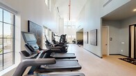 Located at 98 South River Drive in Tempe, Arizona, this fitness center at The Lyssa offers a generously sized, single-floor exercise space designed to support a comprehensive workout experience. The facility's most striking architectural feature is its soaring double-height ceilings, which lend an open, airy atmosphere throughout the space. Abundant natural light streams in through large multi-pane windows positioned along the walls at both lower and upper levels, creating a bright and energizing environment.

The main exercise floor is lined with a row of modern cardio machines, including multiple treadmills and ellipticals, all facing the windows for a pleasant workout backdrop. Adjacent to the cardio area, a well-stocked strength training zone features a multi-station cable machine, weight benches, and a tiered dumbbell rack with a wide range of free weights. A wall-mounted flat-screen TV and built-in storage shelving provide convenience and entertainment, while a water station with a bottle-fill dispenser keeps members hydrated.

A separate glass-enclosed cycling studio houses stationary spin bikes equipped with digital displays, offering a semi-private space for focused indoor cycling sessions. Full-length mirrors are strategically placed to assist with form and to amplify the sense of space. The overall design is clean, modern, and functional, making this fitness center a standout amenity.