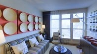 This thoughtfully curated single-floor space makes a strong design statement through its blend of eclectic and contemporary elements. The living area serves as the centerpiece, anchored by a bold red wall-mounted art installation displaying large decorative circular platters — a striking focal point that draws the eye immediately upon entry. The walls are painted in a soft, neutral grey, providing a clean backdrop that allows the colorful furnishings to stand out. A striped multi-color sofa adds a vibrant, casual energy to the seating area, complemented by a sleek modern accent chair in neutral tones. A dark, textured hexagonal accent wall adds depth and visual interest, contrasting elegantly with the lighter surrounding surfaces. Large floor-to-ceiling windows dressed with sheer white curtains and horizontal blinds flood the space with abundant natural light while offering views of the surrounding exterior. A round dark coffee table styled with decorative candle holders and metallic accents grounds the seating arrangement. The overall ambiance is warm, lively, and design-forward, reflecting a carefully curated aesthetic that balances bold artistic choices with comfortable, functional living.