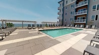 This expansive outdoor amenity space is thoughtfully designed to maximize the stunning waterfront setting it occupies. The centerpiece is a pristine rectangular swimming pool finished in a vivid aquamarine, surrounded by a wide stone-paved sun deck lined with rows of sling lounge chairs — ideal for relaxing and taking in the panoramic open water views that stretch to the horizon.

A pergola structure anchors one end of the terrace, providing a defined architectural focal point while framing the waterscape beyond. The generous paved deck is composed of large-format stone tiles in contrasting light and dark tones, creating an elegant geometric pattern underfoot.

Adjacent to the pool area, a covered outdoor lounge extends along the building's façade, clad in warm wood paneling with floor-to-ceiling glazing. This sheltered zone offers a comfortable seating area with cushioned chairs and a full outdoor dining table, suited for both casual gatherings and al fresco meals. Raised planter beds with lush greenery and seasonal plantings add a natural softness to the hardscape surroundings.

The multi-story contemporary building rises above, featuring balconies, fiber cement siding, and teal-accented window surrounds. Throughout the space, an open, airy ambiance prevails, shaped by abundant natural light, the ever-present water views, and a clean, modern aesthetic.