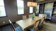 This single-floor space showcases a thoughtfully designed interior that blends rustic charm with functional elegance. The dining area serves as a central focal point, anchored by a substantial parquet-patterned natural wood table surrounded by wood-framed chairs with decorative ikat-style upholstered cushions, adding a touch of color and pattern to the room. A large drum pendant light fixture hangs overhead, casting warm, ambient light across the dining surface. The walls are finished in a rich, deep brown tone that creates an intimate and cozy atmosphere throughout. Two windows fitted with horizontal blinds allow natural light to filter in softly, enhancing the warmth of the space without overpowering it. A built-in shelving and desk unit is seamlessly integrated into one wall, offering dedicated workspace and display storage, complete with open shelving ideal for books, décor, or personal items. The flooring features a textured area rug that adds visual interest and grounds the dining arrangement. Together, these elements create a cohesive, inviting space that balances everyday practicality with refined, casual style.