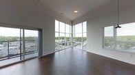 This single-floor space is defined by its striking architectural design, most notably the expansive floor-to-ceiling corner windows that serve as the room's focal point. These large-format windows meet at a corner, flooding the interior with abundant natural light while framing elevated views of the surrounding landscape, including tree canopies and the urban streetscape below. The open main living area features rich, dark hardwood flooring that contrasts cleanly with the crisp white walls and ceiling, contributing to a modern and polished aesthetic. The elevated ceiling height enhances the sense of spaciousness throughout the room. An additional window with blinds is visible on the adjacent wall, further supporting the bright and airy atmosphere. A sliding door or secondary glazed opening on the left side of the space suggests access to a balcony or additional outdoor area. Recessed lighting and a central ceiling fixture provide supplemental illumination. The overall ambiance is contemporary and minimalist, with clean lines, neutral tones, and a layout that maximizes both natural light and the surrounding views.