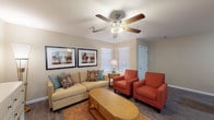 This single-story residence offers a thoughtfully arranged layout with distinct living, sleeping, dining, and kitchen zones visible throughout the floor plan. The living area serves as the heart of the home, featuring wood-look flooring, a ceiling fan with integrated lighting, and windows dressed with white blinds that allow natural light to filter in. Warm neutral walls provide a versatile backdrop, and the space comfortably accommodates a full seating arrangement.

A secondary living or flex area connects seamlessly to the main living space, offering additional room for furniture arrangement and access to natural light through additional windows. The bedroom is generously sized, featuring its own ceiling fan, wood-look flooring, and windows that bring in daylight. It includes access to a spacious walk-in closet outfitted with wrap-around hanging rods on two walls, providing ample storage capacity.

The bathroom is well-equipped with a tiled soaking tub and shower combination featuring safety grab bars, a toilet, an overhead storage cabinet, and a vanity with granite-style countertops. The kitchen area, visible in the floor plan, connects to a dedicated dining space. Throughout the residence, consistent flooring, neutral tones, and ceiling fans contribute to a cohesive and comfortable ambiance.