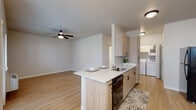 This single-floor residence combines modern design with practical functionality throughout its well-appointed layout. Light wood-style flooring runs continuously across the living spaces, creating a cohesive and airy aesthetic.

The open-concept kitchen is a standout feature, anchored by a peninsula with a built-in sink and white countertops that provide ample prep space and casual seating. Light natural wood cabinetry lines the kitchen walls, offering generous storage, while black appliances — including a range, microwave, and dishwasher — add a contemporary contrast. The kitchen flows seamlessly into the living and dining areas, the latter illuminated by a modern multi-bulb chandelier and a window that brings in natural light.

The living room benefits from ceiling fans with integrated lighting and direct access to a private outdoor area through a glass door. Additional windows ensure the space remains bright throughout the day.

Two bathrooms are finished with distinctive patterned tile flooring that adds character, paired with light wood vanities and white countertops. One bathroom features a walk-in shower, while the other includes a toilet and vanity area.

A dedicated in-unit laundry closet houses a full-sized side-by-side washer and dryer, adding significant convenience. Additional rooms with light wood flooring and neutral walls offer flexible living or sleeping spaces, each with natural light from windows.