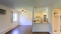 This single-floor space showcases a thoughtfully designed open-concept layout that connects the living area and kitchen, creating a cohesive and functional environment. The kitchen is well-appointed with crisp white cabinetry, granite countertops, and stainless steel appliances, including a full-size refrigerator. A peninsula counter extends into the space, offering additional prep area and casual seating. Recessed lighting illuminates the kitchen, highlighting the clean, contemporary aesthetic. The living area benefits from a ceiling fan and a window that allows natural light to filter in, contributing to the bright, airy atmosphere throughout. Rich hardwood-style flooring runs continuously across both areas, grounding the space with warmth and visual continuity. White walls and neutral tones create a clean backdrop, making the space feel open and inviting. The structural column between the kitchen and living area adds a subtle architectural detail while maintaining the open flow of the layout. Overall, the space presents a practical yet polished environment suited for everyday living.