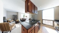 This single-floor space showcases an open-concept layout that integrates the kitchen and living area into one cohesive environment. The kitchen is anchored by a peninsula with dark granite countertops, rich dark-wood cabinetry both above and below, and a stainless steel double-basin sink. Overhead cabinetry suspended from the ceiling defines the kitchen boundary without obstructing sightlines, maintaining the open flow between spaces. A decorative tile backsplash adds a refined visual detail behind the sink area. The kitchen connects directly to the living area, which features neutral-toned carpet, a dark wood coffee table, and upholstered seating arranged around the space. Large windows along the perimeter fill both areas with abundant natural light, creating a bright and airy atmosphere throughout. Light tile flooring in the kitchen transitions smoothly into carpeting in the living area. The overall design balances functionality with contemporary style, incorporating warm wood tones against neutral walls and dark stone surfaces for a polished, inviting aesthetic.