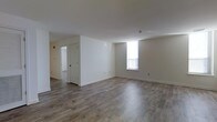 This single-floor space presents a clean, minimalist aesthetic throughout its open layout. The main living area is finished with wood-style laminate flooring in a warm gray-brown tone, lending a contemporary feel underfoot. Crisp white walls and white trim create a bright, neutral backdrop that allows for versatile furnishing and décor options. A window positioned along one wall draws in natural light, brightening the space and adding an airy quality to the room. The ceiling is smooth and standard height, contributing to the open, uncluttered atmosphere. A hallway visible from the main living area leads to additional rooms, with a white paneled door offering access to a separate space — suggesting a functional and well-organized floor plan. The overall design is straightforward and practical, with clean lines and a move-in-ready finish. The consistent flooring flows throughout, tying the spaces together cohesively. The neutral palette and simple detailing make this a versatile space suitable for a range of uses and personal styles.