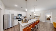 This single-story residence in West Columbia, SC offers a thoughtfully designed layout across three bedrooms and two bathrooms. The open-concept living area serves as the heart of the home, where the kitchen, dining room, and living room flow seamlessly together. The kitchen is appointed with crisp white shaker cabinetry, granite countertops, stainless steel appliances, and a large island with bar seating illuminated by pendant lighting — ideal for both cooking and casual entertaining. Wood-look flooring extends throughout the main living areas, adding warmth and continuity.

The dining area sits adjacent to the kitchen, while the living room features a decorative column detail and a window that brings in natural light. A carpeted hallway connects the bedroom wing to the main living spaces. The primary bedroom offers a comfortable, cozy atmosphere with carpet flooring and a window overlooking the outdoors. The bathrooms include a tiled tub-and-shower combination. A dedicated laundry room houses a full-size washer and dryer with built-in wire shelving for added storage. The neutral color palette and clean architectural lines throughout lend the space a modern, move-in-ready appeal.